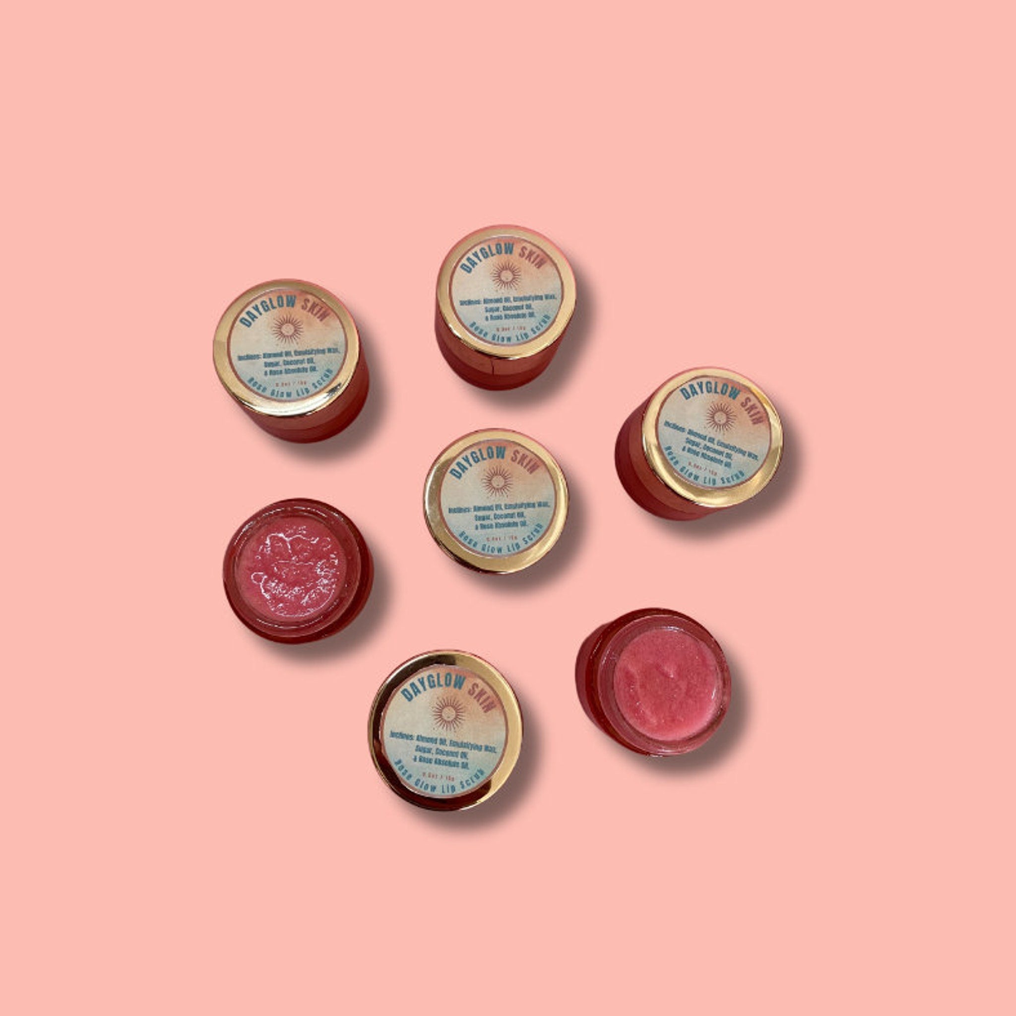 Lip scrub jars, red and gold containers, DAYGLOW SKIN branding visible on the lids, on a pink background