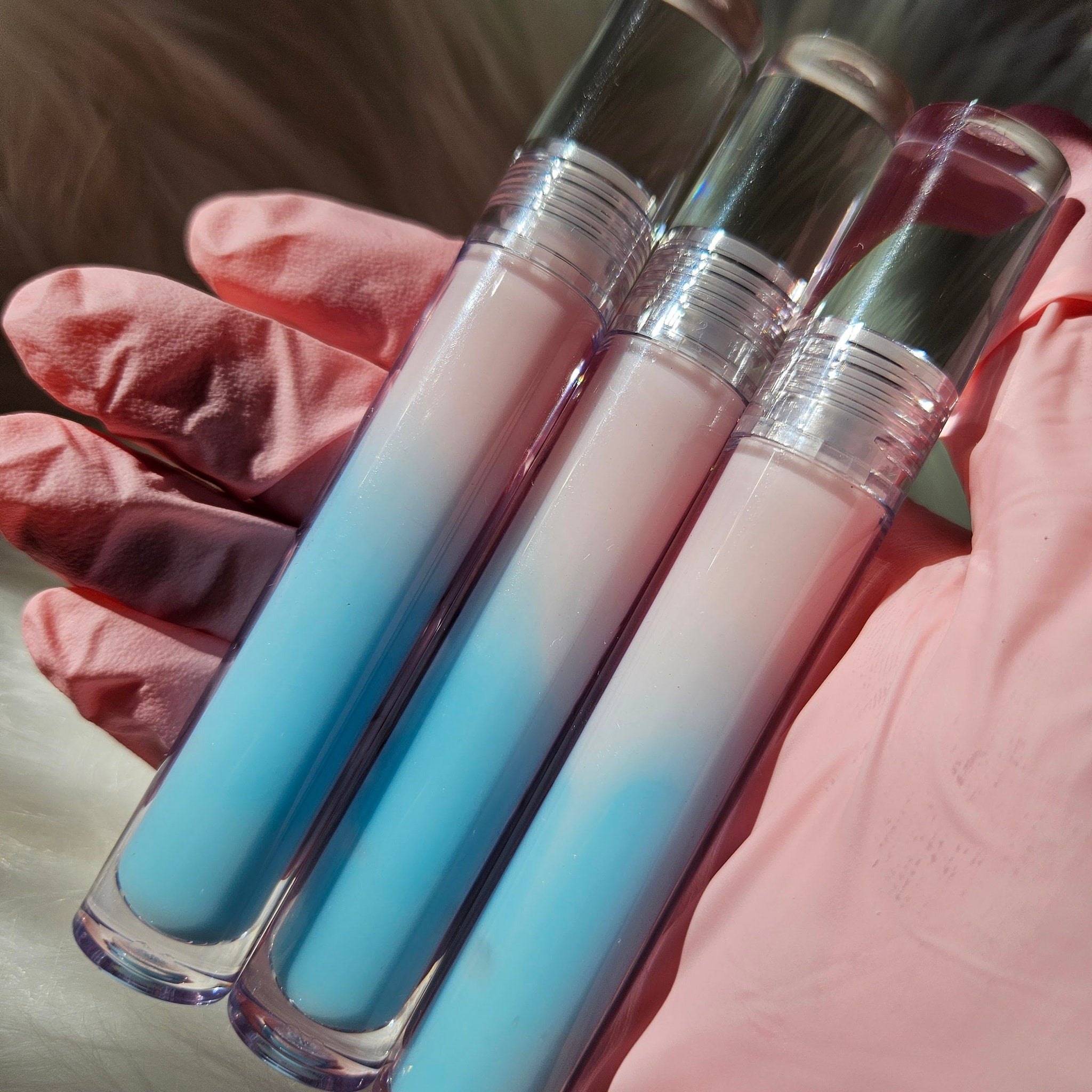 Lip gloss tubes with clear plastic, blue and pink gradient, held in pink gloved hand