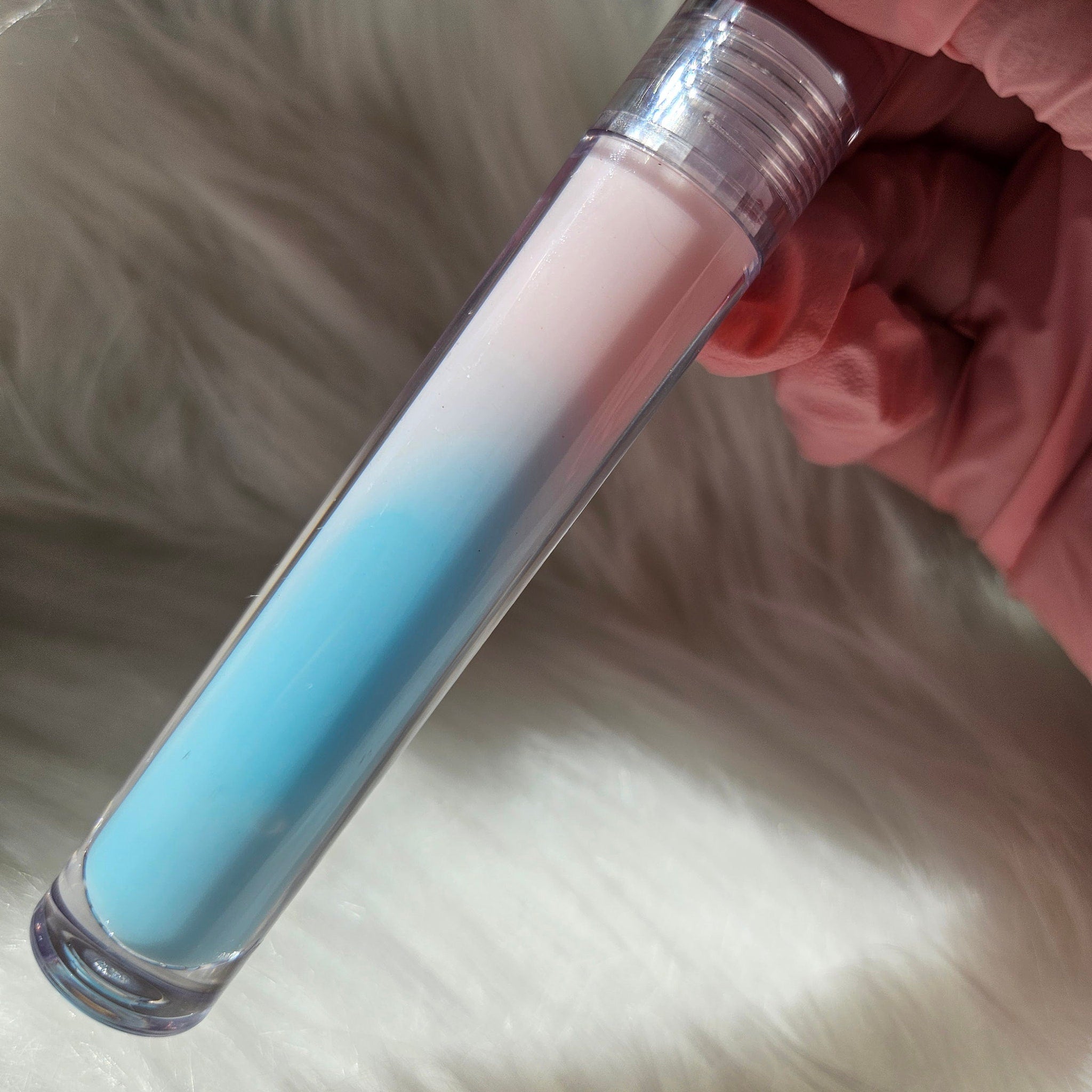 Lip gloss tube with pink and blue gradient, clear plastic, held by pink-gloved hand