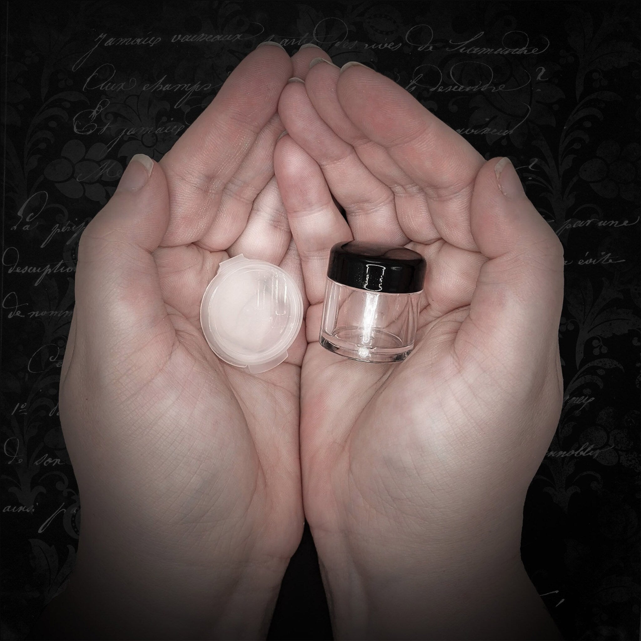 Small clear jar with black lid and white lid, held in hands against a dark background