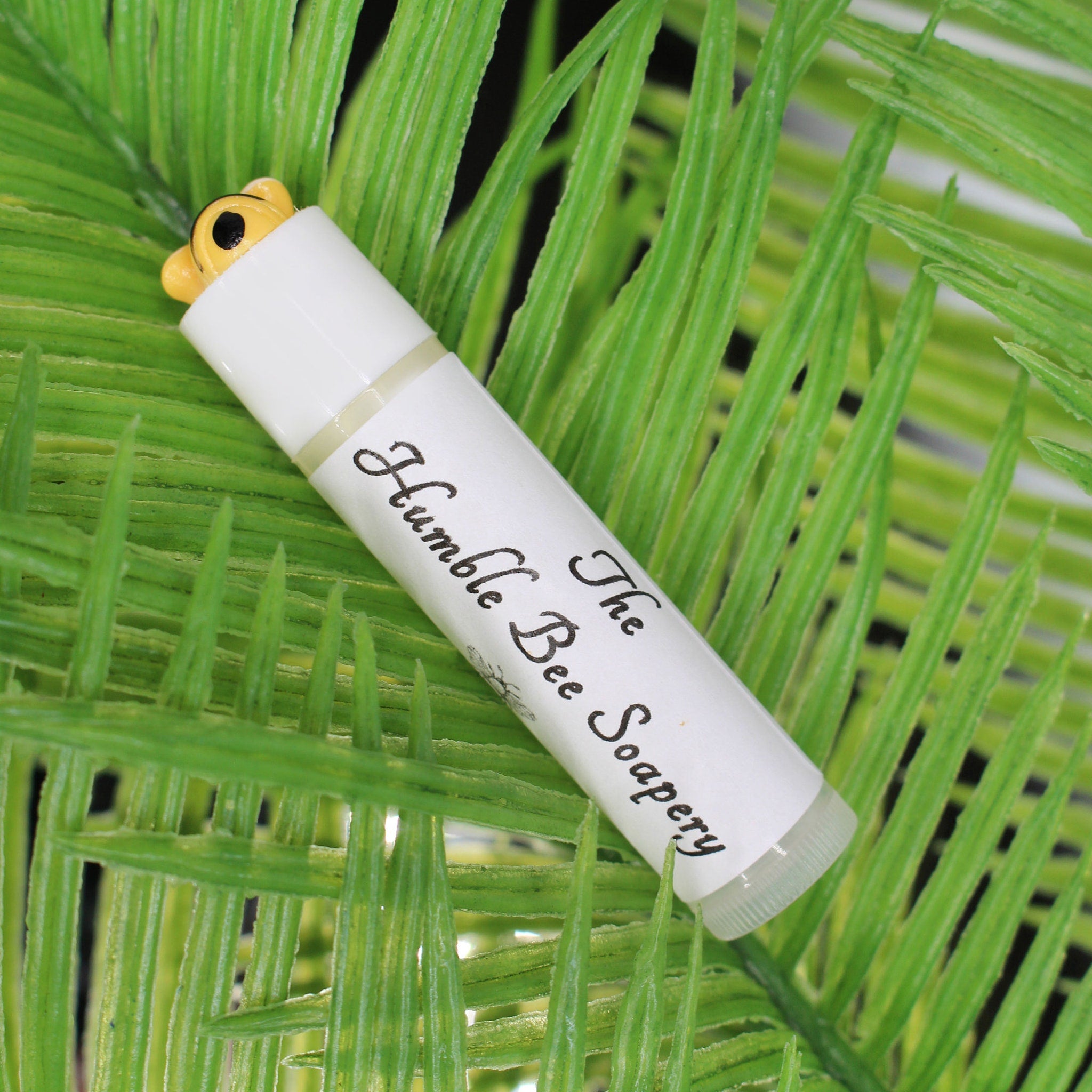 Lip balm tube with white label, yellow bee top, and The Humble Bee Soapery text on green leaf background