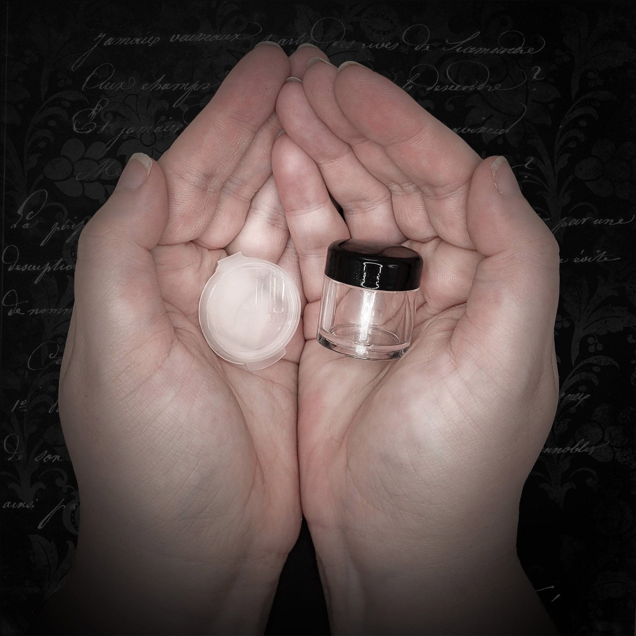 Small clear jar with black lid and white lid, held in cupped hands against a dark floral background