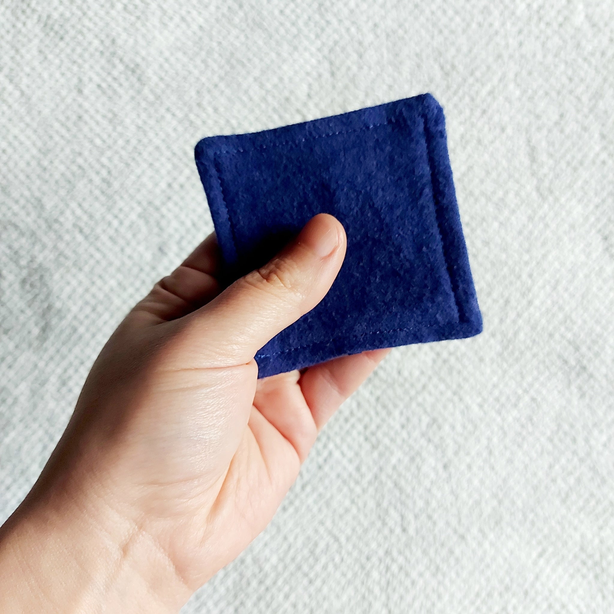 Reusable facial pad, navy blue felt square held in hand, showing stitched edge detail