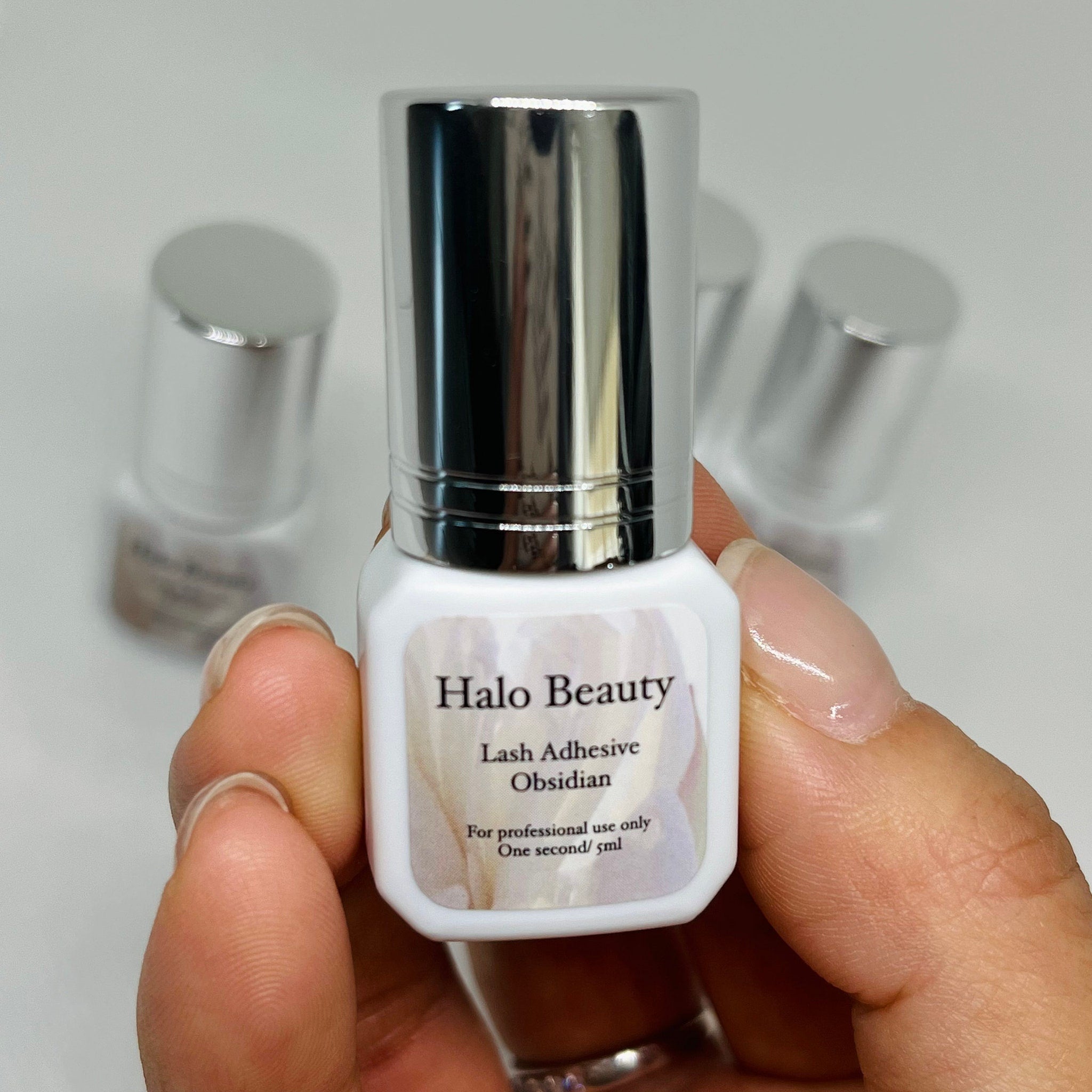 Lash adhesive bottle with silver cap, white label, held by hand, Halo Beauty brand name visible