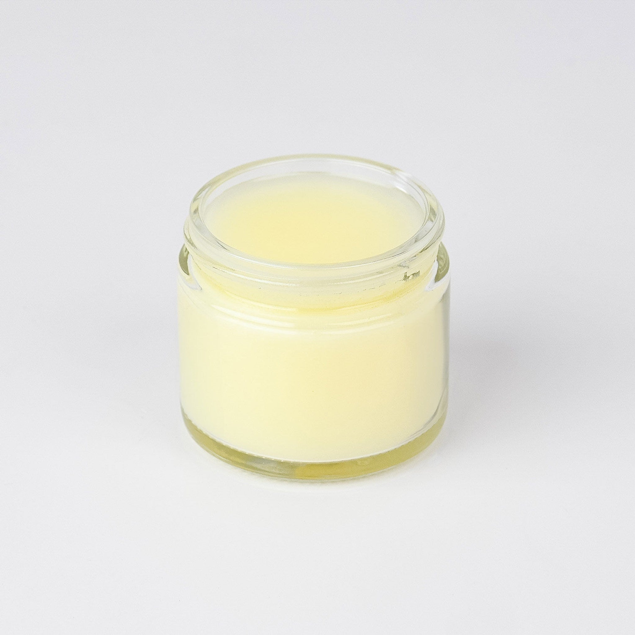 Lip balm in a clear glass jar with pale yellow balm inside, on white background