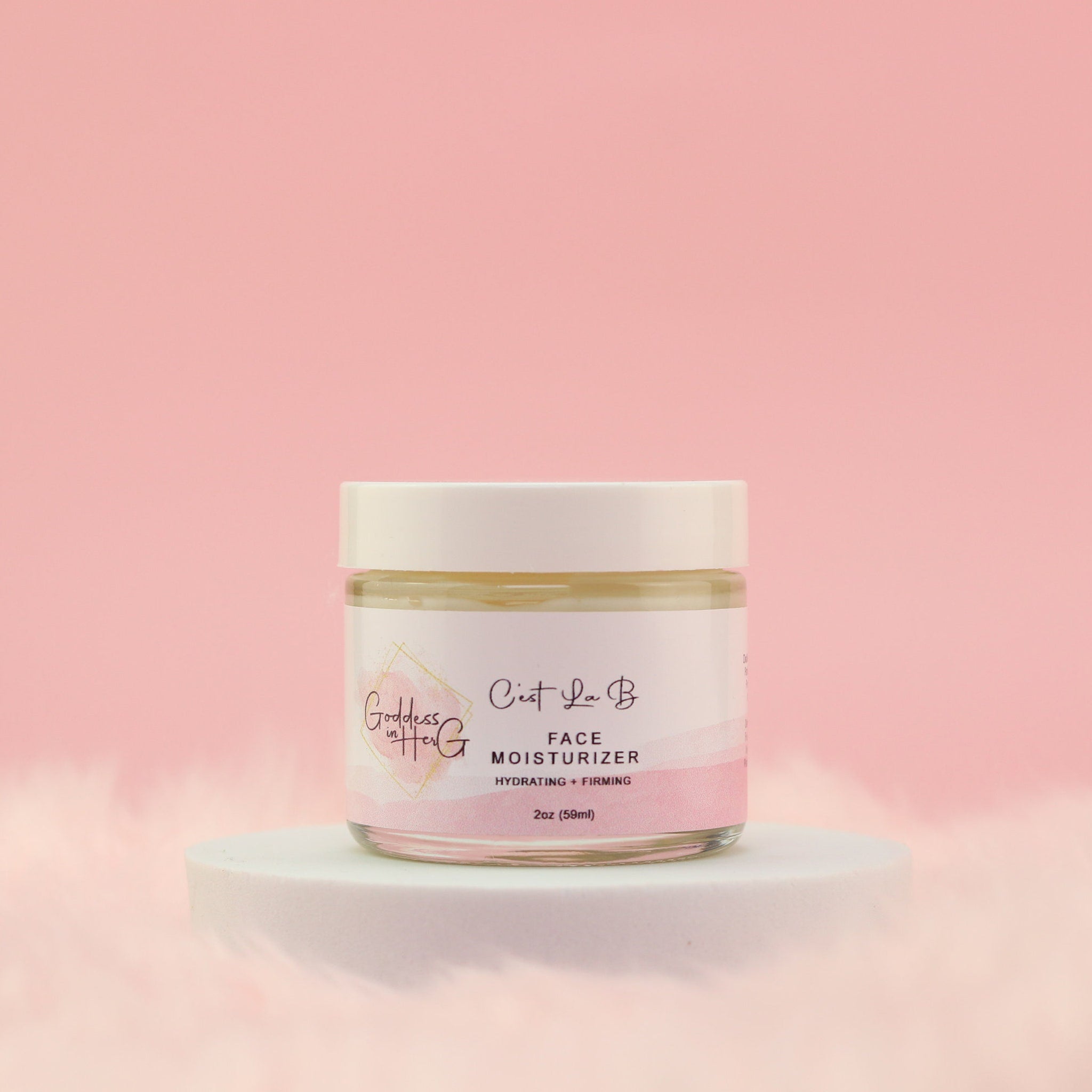 Face moisturizer jar with white lid, glass jar, and pink label on white pedestal