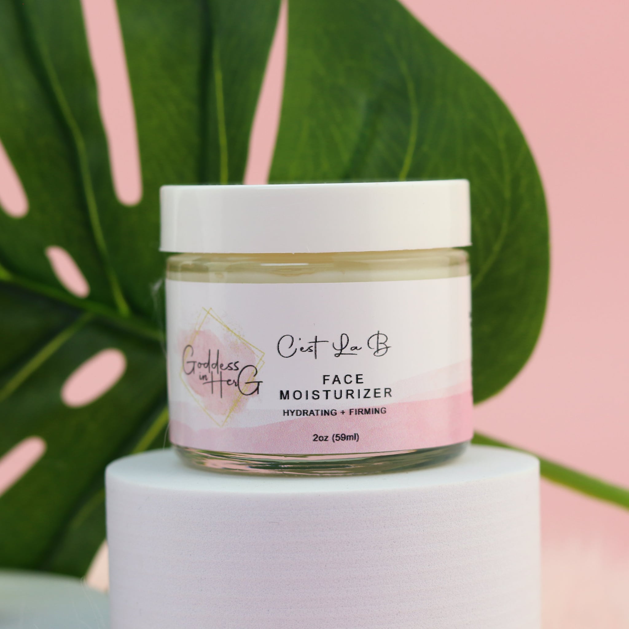 Face moisturizer jar with white lid, pink and white label, and Goddess in Her G branding on a white stand