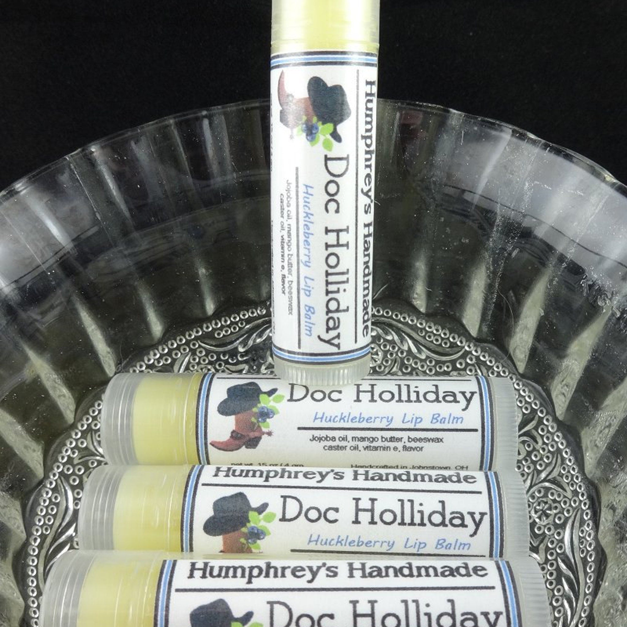Lip balm tubes with white labels, blue accents, and "Doc Holliday" text, stacked in a glass dish