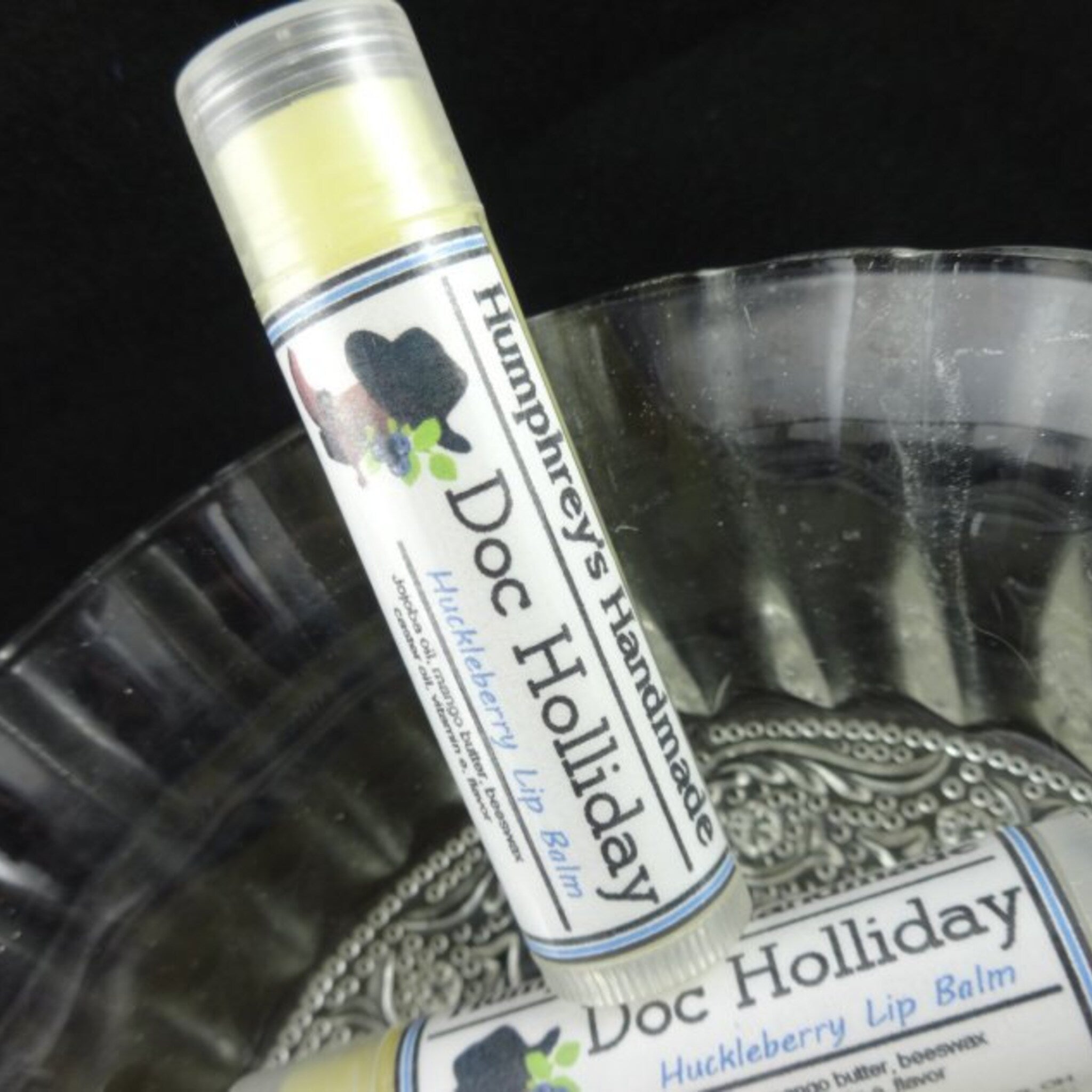 Lip balm tube with yellow balm, clear plastic, and Doc Holliday label on a decorative glass dish