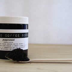Coffee scrub jar with black and white label, white lid, and dark granules on wooden spoon