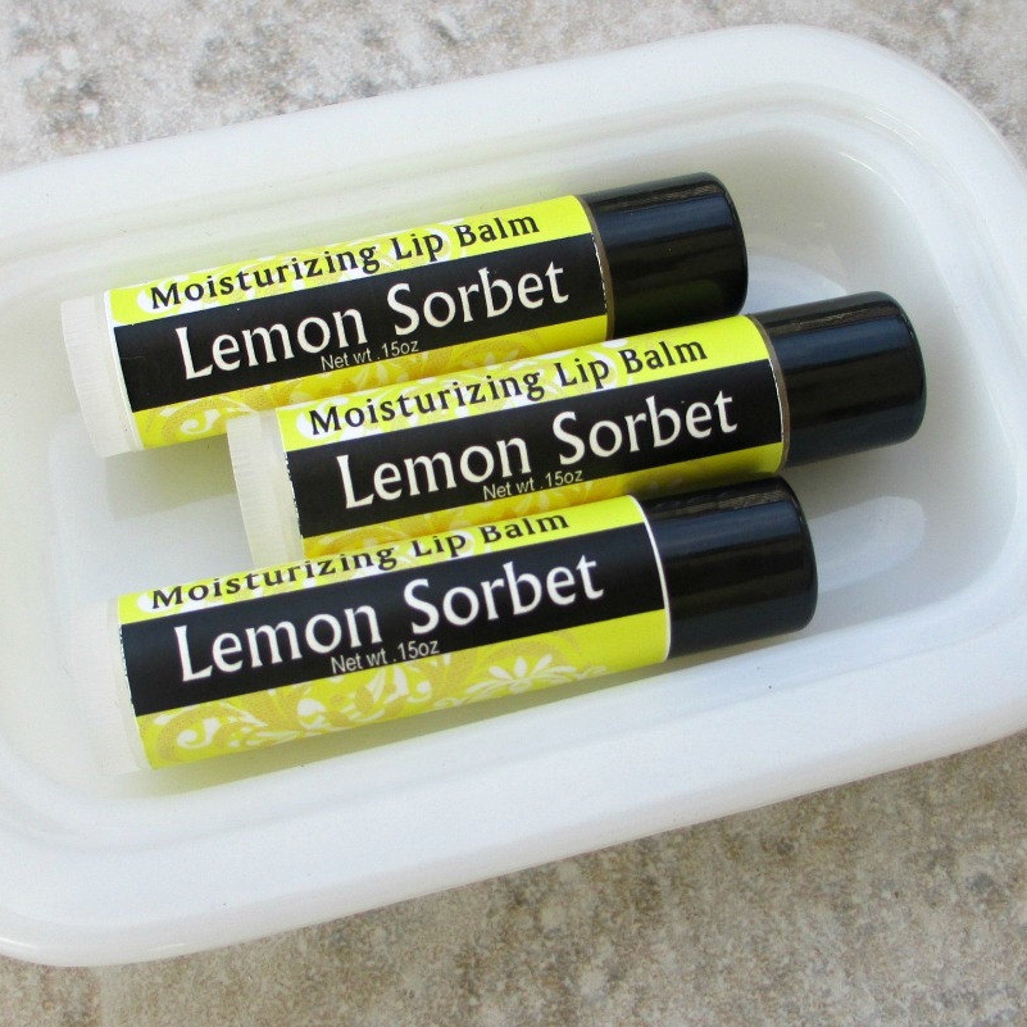 Lip balm tubes with yellow and black labels, black caps, in a white dish, "Lemon Sorbet" text