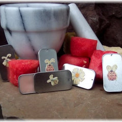 Lip balm tins with floral illustrations, silver and black metal, next to watermelon