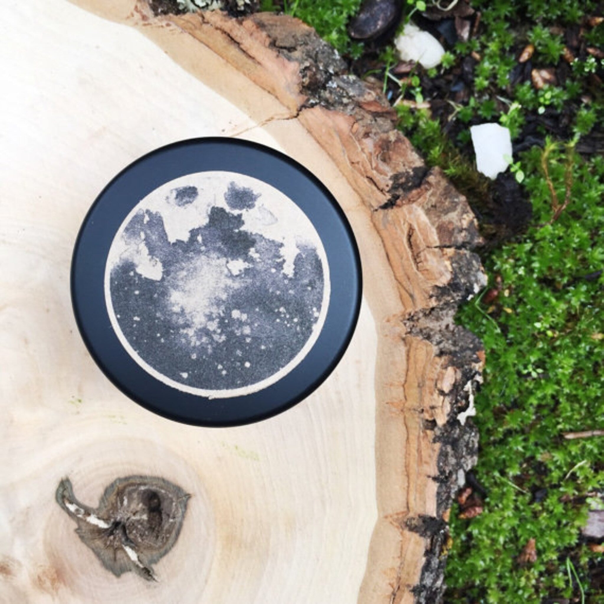 Black round container with moon design on wood surface and green moss background