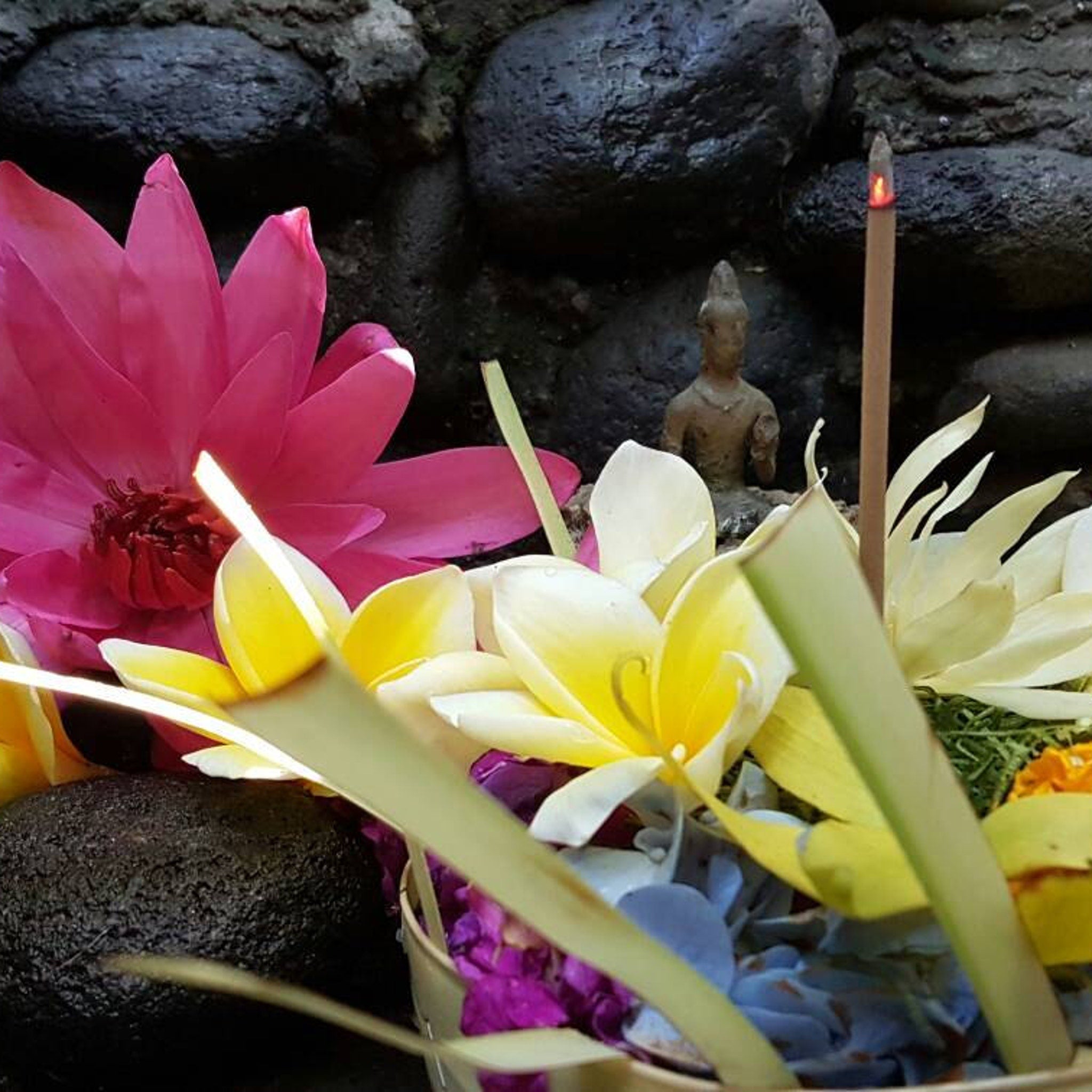 Pink and yellow flowers with burning incense stick and small statue in front of dark stone background